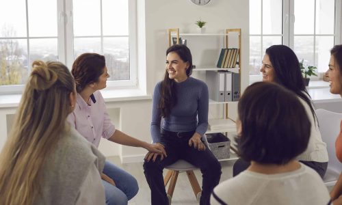 Happy thankful young women sitting in circle, smiling, talking and sharing positivity in group meeting or therapy session with coach. Female community, support and overcoming problems together concept